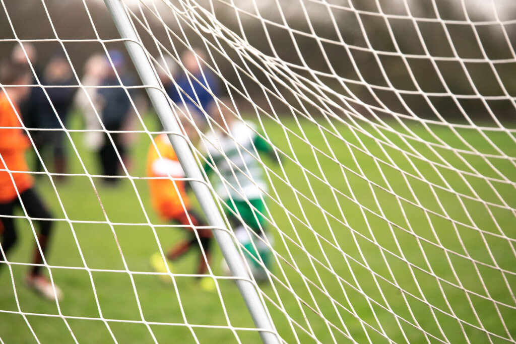 boys playing football match from behind goal net with intentional blur of identities, representing sports clubs