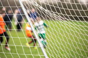boys playing football match from behind goal net with intentional blur of identities, representing sports clubs